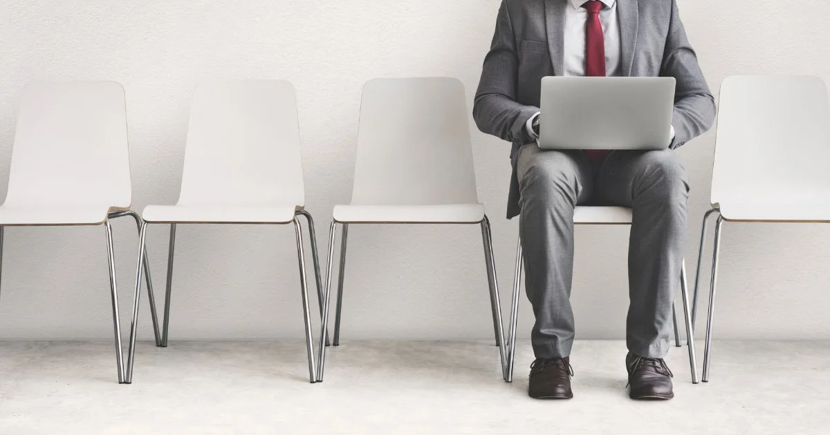 Businessman in a suit using a laptop while seated among empty white chairs in a waiting area.